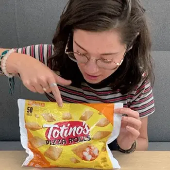 A girl with glasses sitting at a table pointing at a bag of Totino's Pizza Rolls. - Link to social post