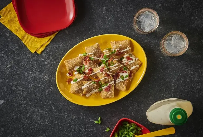 Two glasses of water, napkins, a bottle of ranch & a long yellow bowl in the centre of a table containing Totino's Cheesy Bacon Ranch Pizza Rolls recipe.