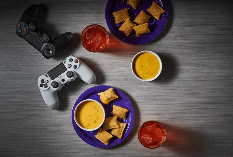 A table with Playstation controllers, two glasses of orange soda & plates with Totino's Pizza Rolls & a side of Zesty Cheese Pizza Dipping Sauce.
