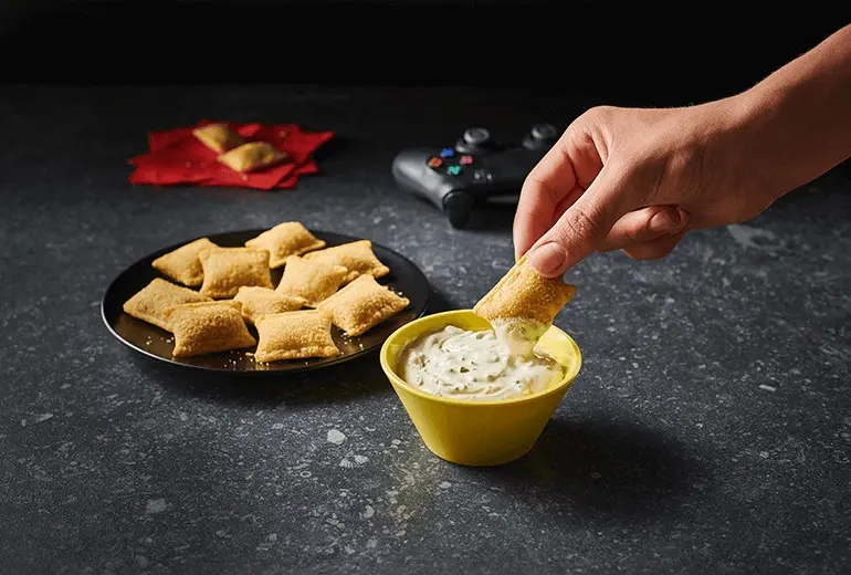 A hand dipping a Totino's Pizza Roll into Totino's Cold Jalapeno Popper Dip recipe alongisde a plate of Pizza Rolls and a black game controller.
