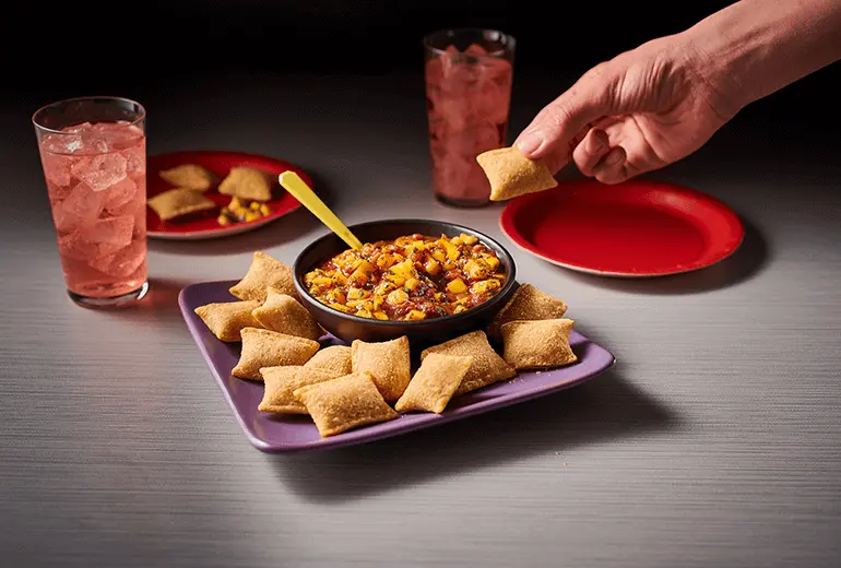 A hand dipping Totino's Pizza Rolls into Totino's Easy Mango Salsa Recipe, surrounded by more Pizza Rolls & two glasses of red soda with ice.