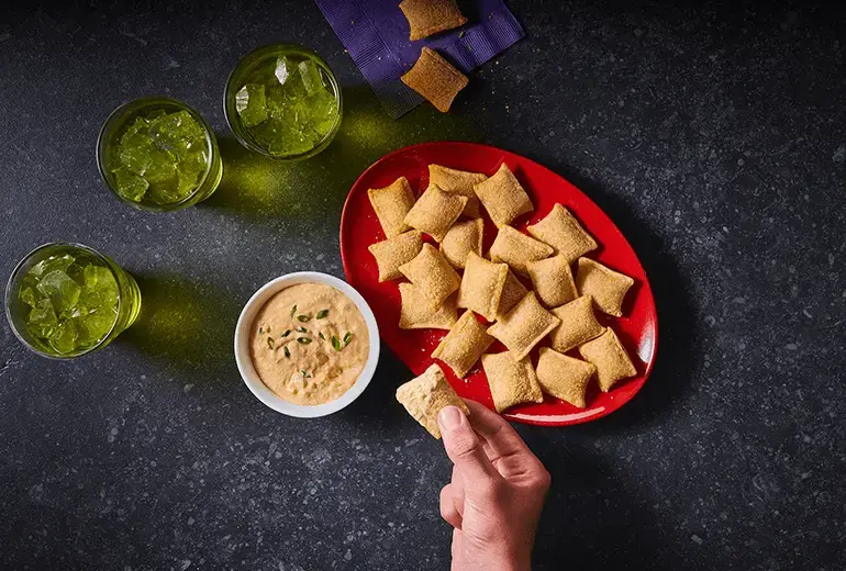 Three glasses of water, napkins, a bowl of dip, & a long red bowl in the centre of a table containing Totino's Cheesy Bacon Ranch Pizza Rolls recipe.