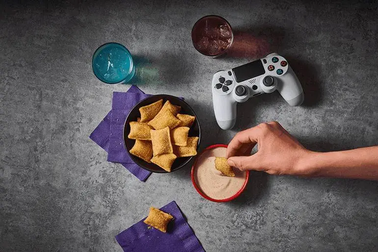 A hand dipping Totino's Pizza Rolls into Totino's Ridiculously Good Ranch Sauce Dip recipe accompanied by a bowl of Pizza Rolls, game controllers & soda.
