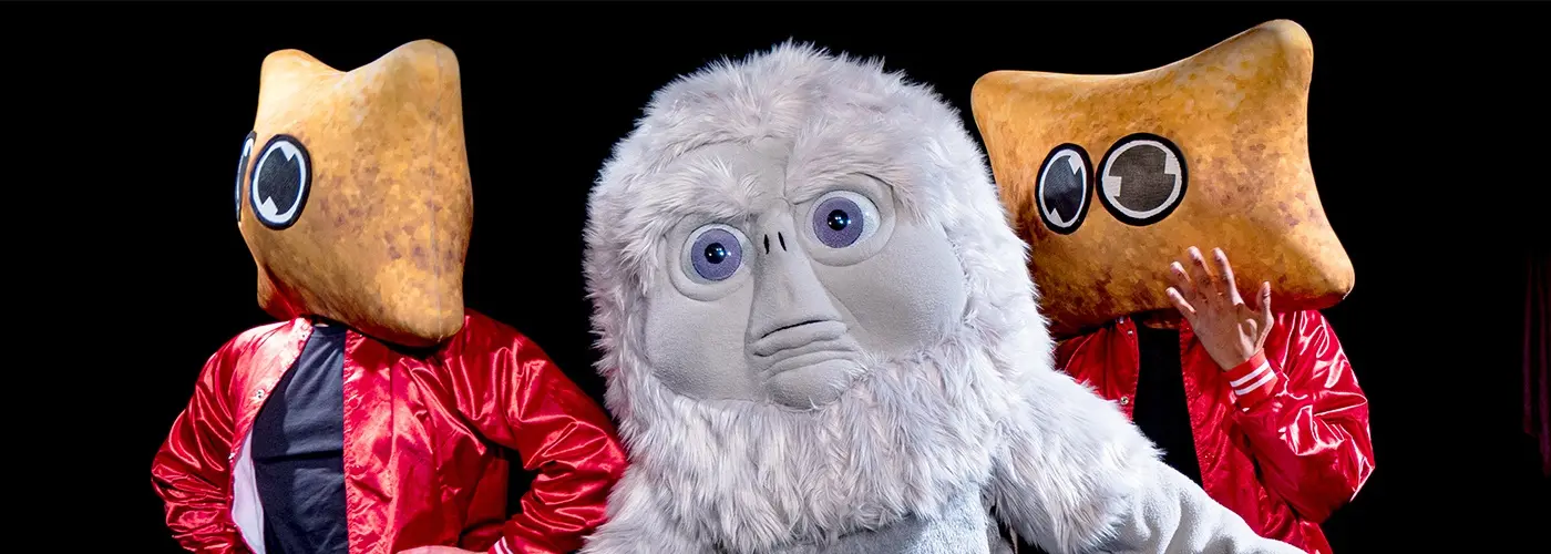 Two people in red satin jackets wearing large, golden, snack-shaped mascot heads stand on either side of a fluffy white mascot character against a dark background, with a blurred rectangle centered over the middle figure.
