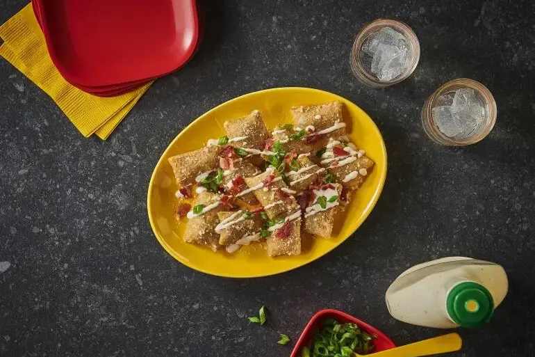 A plate of fried ravioli topped with diced tomatoes, green onions, and drizzled with a creamy sauce is placed on a table. Nearby are two glasses filled with ice, a bottle of sauce, a bowl of chopped green onions, and stacked plates with napkins.